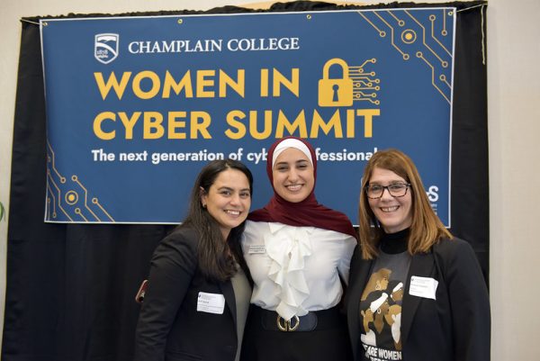 Three women -two brunettes, and one in a maroon hijab- pose in front of a blue banner at the champlain college women in cybersecurity summit.